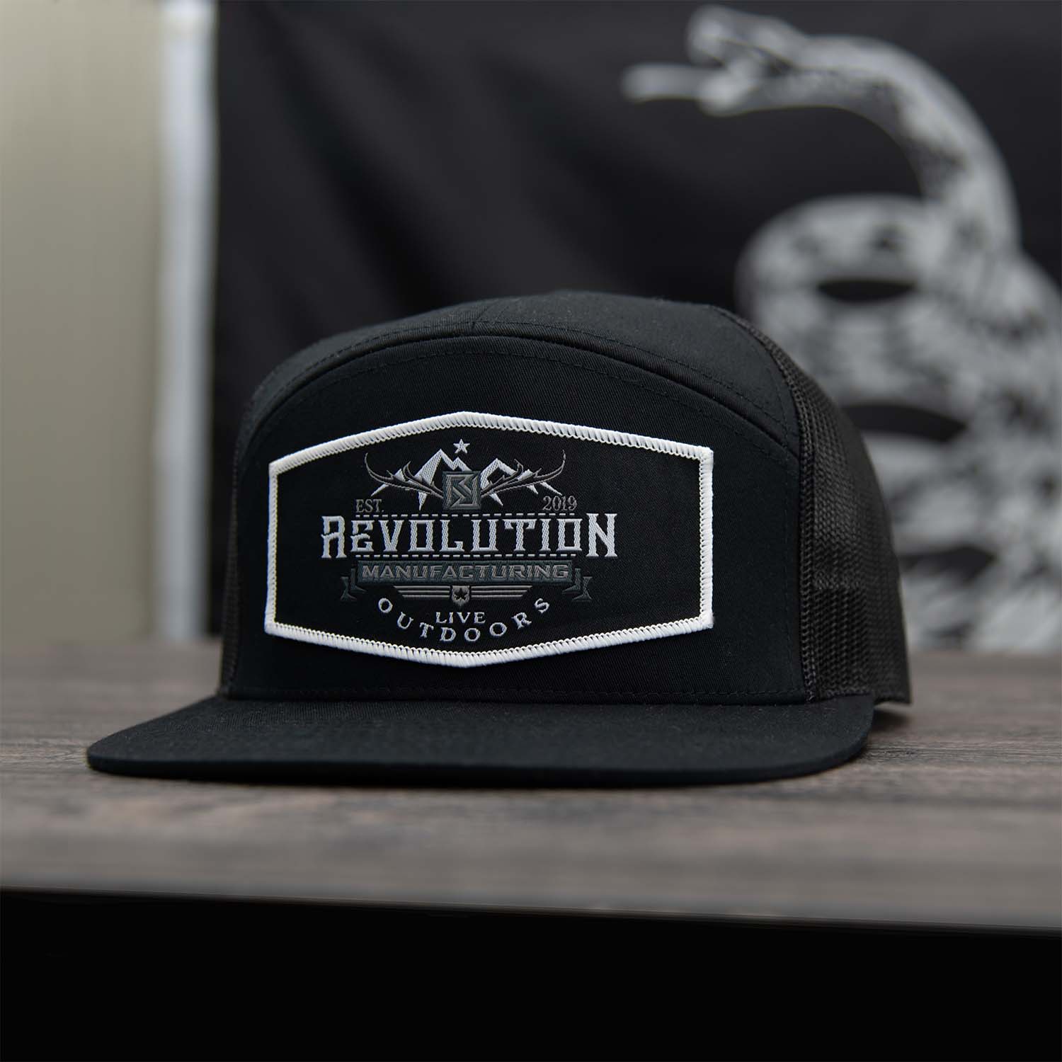 All black 7 panel flat bill trucker hat with Revolution Mfg Live Outdoors woven black patch with white border