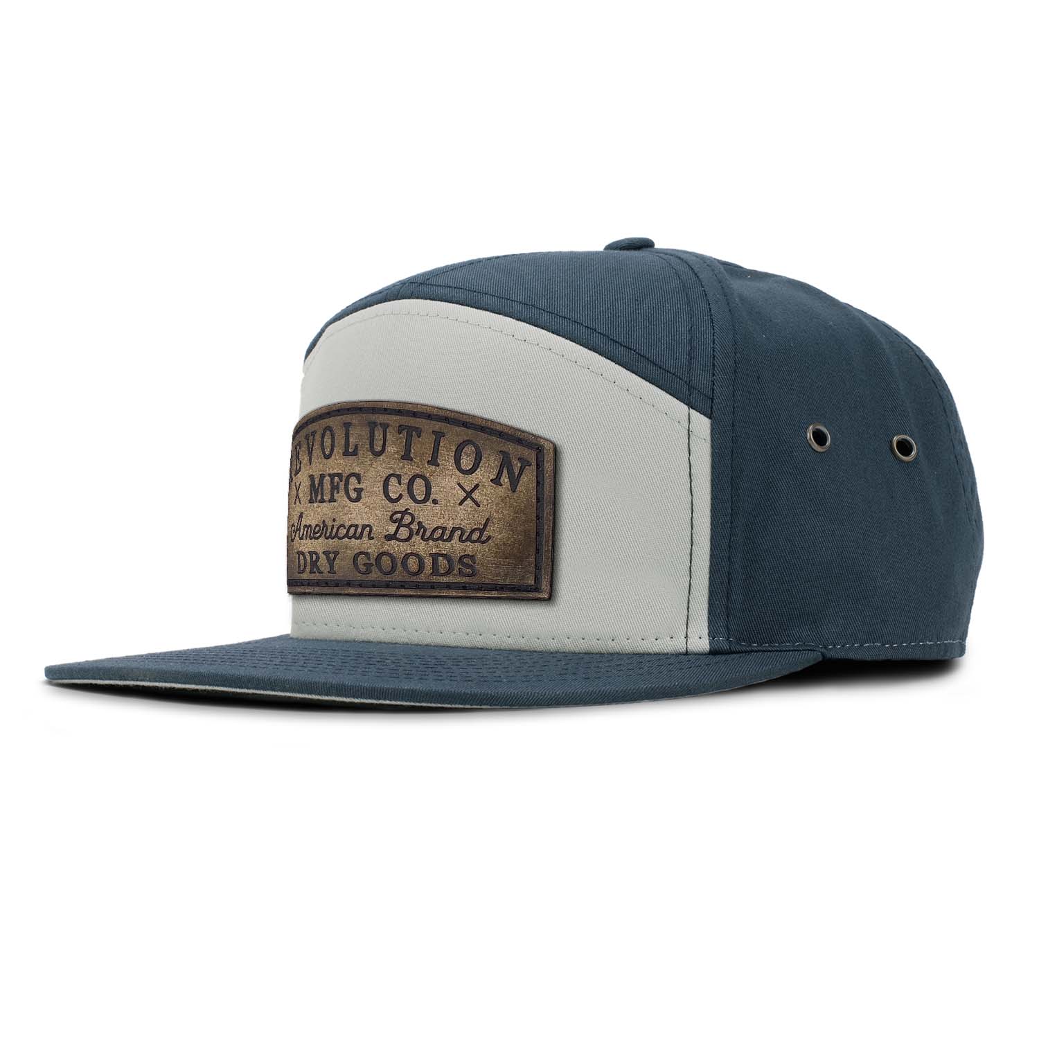 A pigeon gray and navy 7 panel style hat featuring an adjustable leather strapback with brass clasp closure and brass eyelets with our full grain leather vintage Dry Goods patch on the seamless front panel